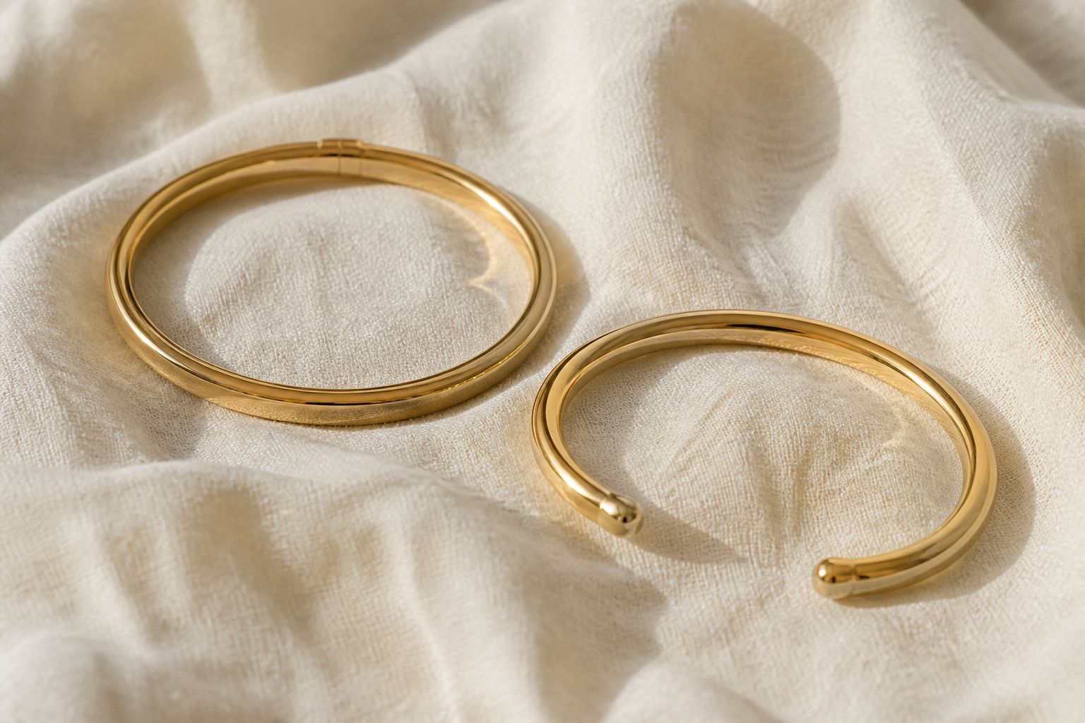A closed gold bangle beside an open gold cuff on linen